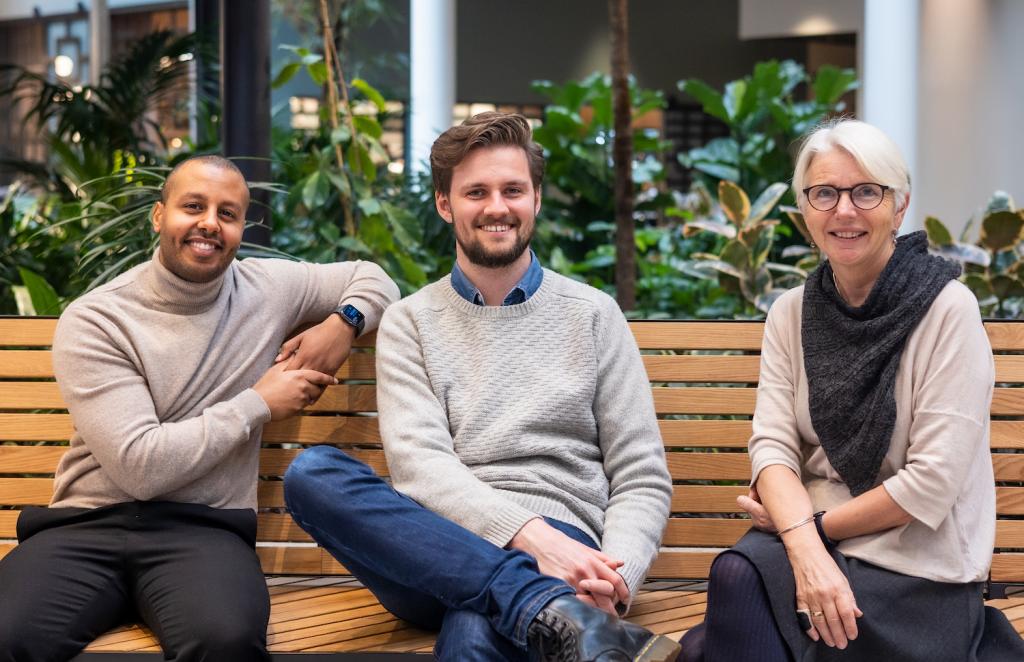 Tre personer fra Ressursenteret, Hermon, Jens og Tone, sitter på en benk og ser mot kameraet med et smil