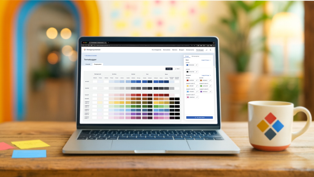 Åpen bærbar pc som står på et trebord. Ved siden av står en kaffekopp med logoen til Designsystemet. 