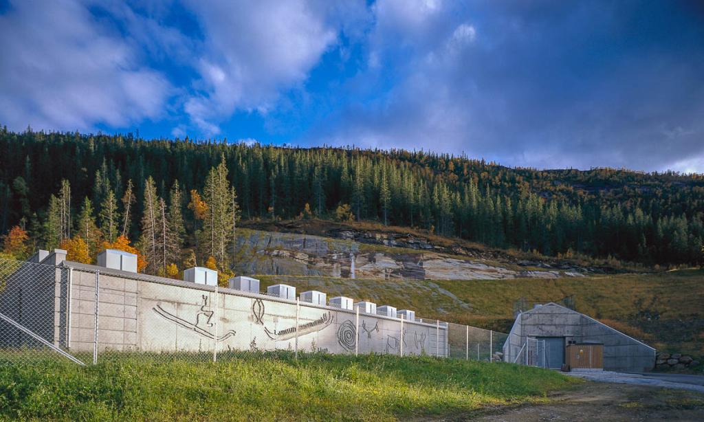 Betongbygning og lukket tunnellinngang i et fjell med granskog på toppen og blå himmel. Fotografi av arkivverkets lagringsanlegg i Mo i Rana.